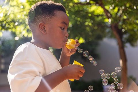 African american boy blowing bubbles in sunlit backyard with yellow wand