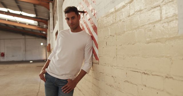 Young man standing inside an empty warehouse, leaning against a white brick wall. He wears a white long-sleeved shirt and jeans. This can be used in contexts related to casual fashion, modern urban lifestyle, introspection, and relaxed atmosphere.