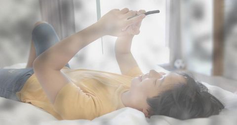 Asian man lying on bed holding tablet above face scrolling in bright bedroom