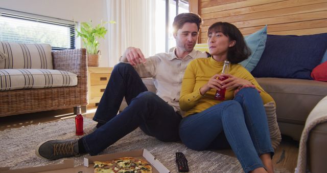 Couple sitting on floor in comfortable living room, enjoying pizza and drinks together. Great for themes of home life, relaxation, domestic bliss, quality time, casual lifestyle, and intimate moments. Ideal for advertisements or articles related to couples, indoor activities, cozy interiors, and enjoying weekend downtime.