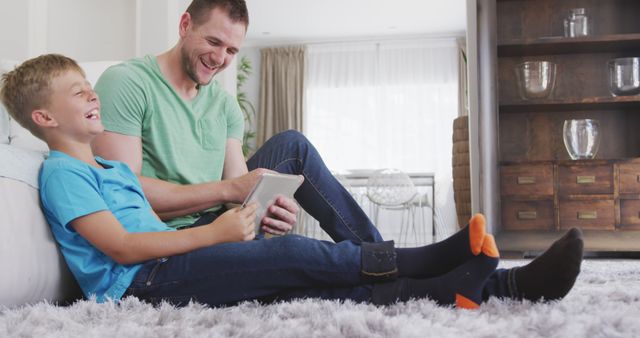 Happy caucasian father and son sitting on floor, using tablet and laughing. Lifestyle, domestic life, communication, family, and togetherness.