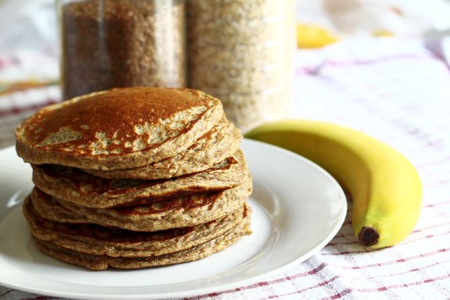 Stack of homemade whole grain pancakes on white plate with a ripe banana on a red-and-white checkered cloth backdrop. Ideal for use in food blogs, healthy eating articles, breakfast recipe features, or diet and nutrition promotions.