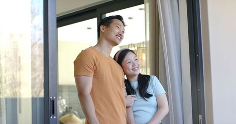 Asian couple standing on balcony smiling and looking through sliding glass door at home