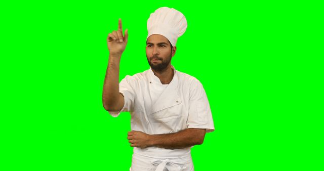 A professional chef in full uniform is pointing upwards against a green screen background. This image is ideal for cooking shows, recipe apps, culinary tutorials, or restaurant promotions. The green screen allows easy background replacement for various creative projects.