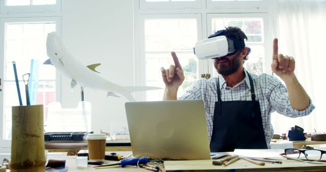 A man wearing a virtual reality headset is working on 3D modeling at his desk, using a laptop and surrounded by various tools in a well-lit room. This scene can be useful for illustrating stories related to modern technology, innovation in design, immersive experiences, and creative workspaces.