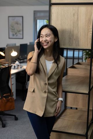 Asian businesswoman leaning on shelf smiling while talking on smartphone in modern open-plan office