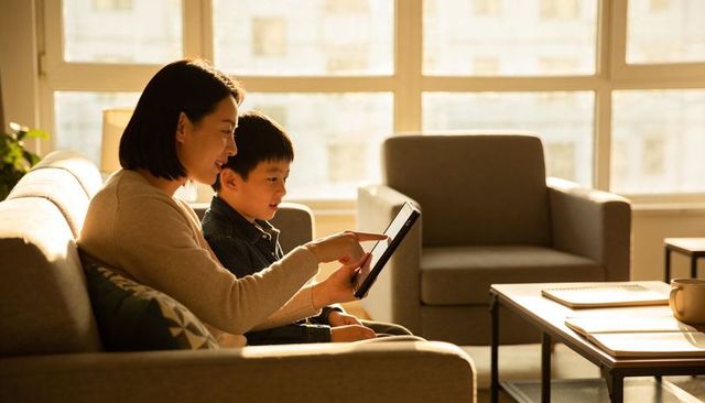 Mother teaching son with tablet on sunlit living room sofa for family learning and bonding