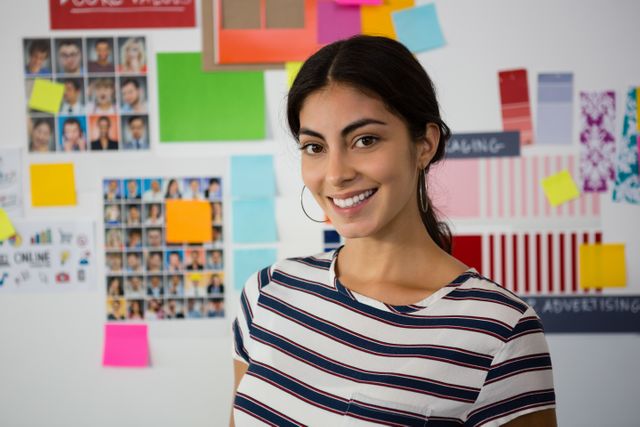 Young woman standing in a creative office environment, smiling confidently. The background features colorful sticky notes and various images, suggesting a brainstorming or planning session. Ideal for use in business, creativity, and professional development contexts, as well as for promoting office culture and teamwork.