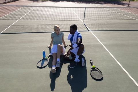 Diverse tennis partners resting and chatting on sunlit hardcourt