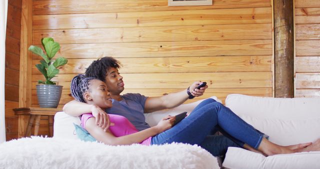 Happy african american couple watching tv, sitting on sofa in log cabin, slow motion. Domestic life, countryside and nature concept.