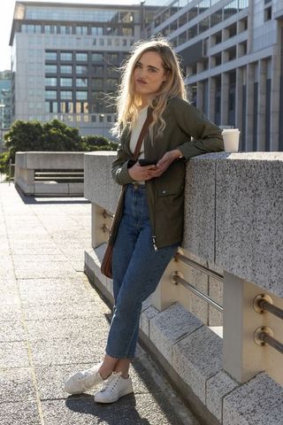 Young woman leaning on rooftop balustrade using smartphone while coffee cup rests on ledge