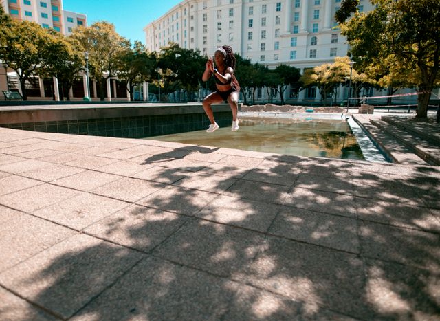 African American woman wearing earphones, jumping during exercise in urban park. Ideal for promoting healthy lifestyle, fitness programs, outdoor activities, and urban living. Suitable for use in fitness blogs, health magazines, and advertisements for sportswear or earphones.