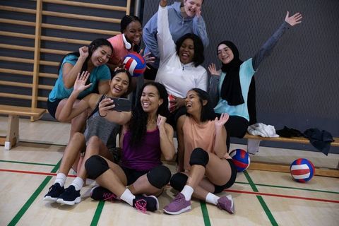 Diverse women's volleyball team celebrating together in gymnasium taking joyful selfie