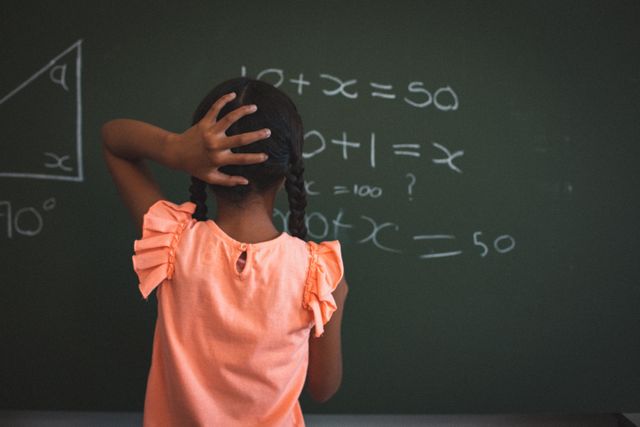 This image shows a young schoolgirl standing in front of a chalkboard filled with math problems, holding her head in confusion. Ideal for educational content, articles on childhood learning challenges, math tutoring services, and classroom environment visuals.