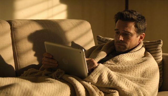 Middle-aged man wrapped in beige knit blanket using tablet on sofa in warm golden light