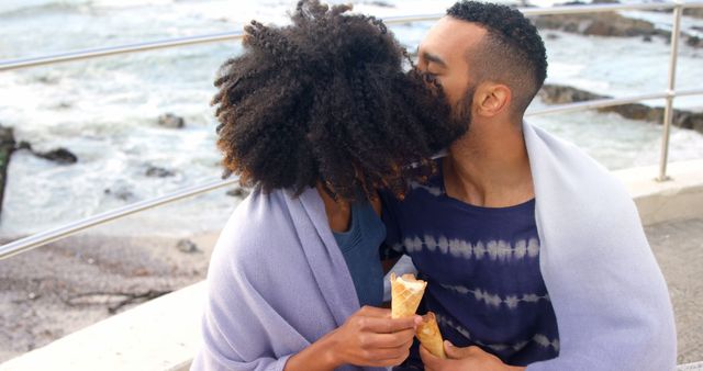 Romantic diverse couple with blankets on backs and ice creams embracing and kissing on sunny beach. Summer, vacation, romance, love, relationship, free time and lifestyle, unaltered.