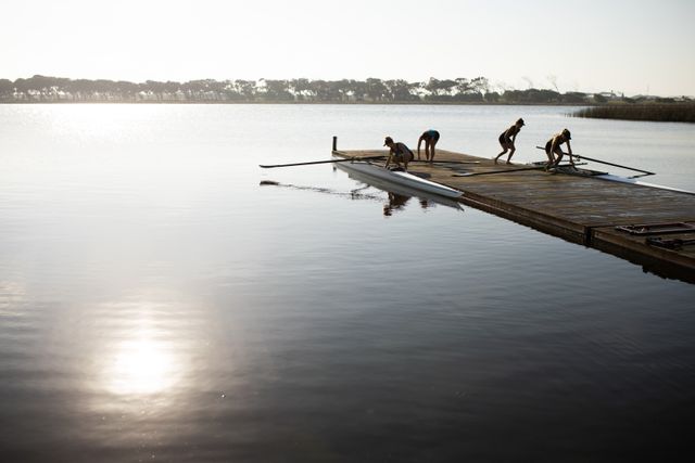 Rowing team of four Caucasian women preparing their racing shell on a jetty at sunrise. Ideal for use in articles or advertisements about teamwork, fitness, water sports, morning routines, and outdoor activities.