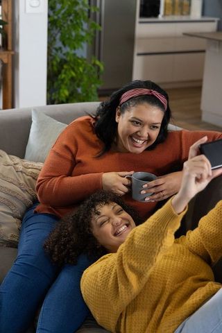 African american mother and teen daughter laughing and relaxing on sofa with smartphone