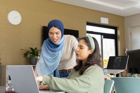 Female colleagues collaborating at laptop in modern office with woman wearing blue hijab