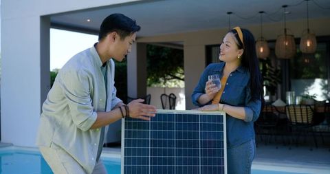 Asian couple examining solar panel at poolside patio, discussing sustainable home energy