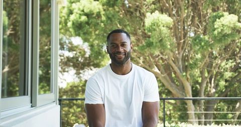 Smiling man relaxing on sunlit balcony wearing white t-shirt with lush garden background