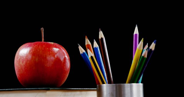 A bright red apple sits next to a container filled with colorful pencils against a dark background, with copy space. These items are often associated with education and can symbolize learning or back-to-school themes.