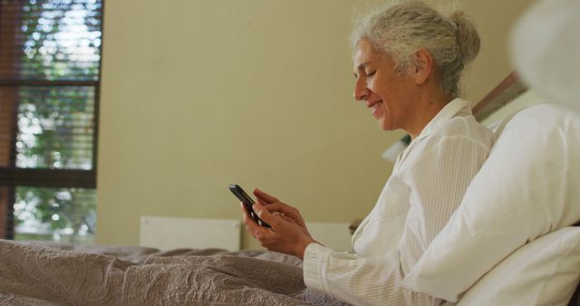 Senior woman smiling while using smartphone in bed on sunny morning - Download Free Stock Images Pikwizard.com