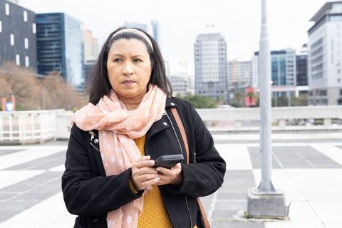 Mature woman checking smartphone in urban plaza wearing peach scarf and shoulder bag