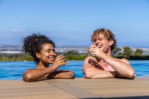Couple relaxing with drinks and laughing at infinity pool overlooking coastal horizon