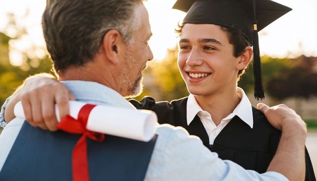 Father hugging graduate son holding diploma with red ribbon at golden hour