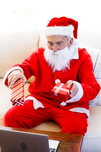 Front view of an old caucasian man wearing a santa clause costume holding out a christmas gift in front of him with each hand. he is sitting on the couch in the living room.
