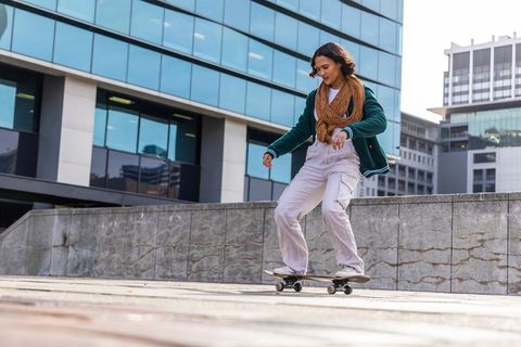 Young woman skateboarding in urban plaza balancing on skateboard near modern glass offices
