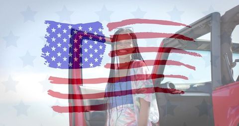 Smiling woman with braids by red car wearing sunglasses and printed top with american flag overlay