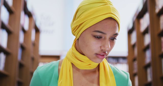 African woman wearing yellow hijab is surrounded by bookshelves in library, focused on reading. Ideal for illustrating themes related to education, cultural diversity, and student life. Good for use in educational materials, diversity initiatives, and library promotions.