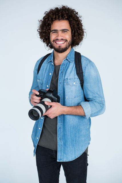 Young male photographer with curly hair wearing a denim shirt, holding a professional camera and smiling in a studio. Ideal for use in articles about photography, creative professions, hobbies, or promotional materials for photography courses and workshops.