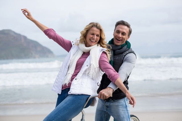 Mature couple enjoying a joyful bicycle ride on the beach with ocean waves in the background. Ideal for use in advertisements, travel brochures, lifestyle blogs, and content promoting healthy living, outdoor activities, and romantic getaways.