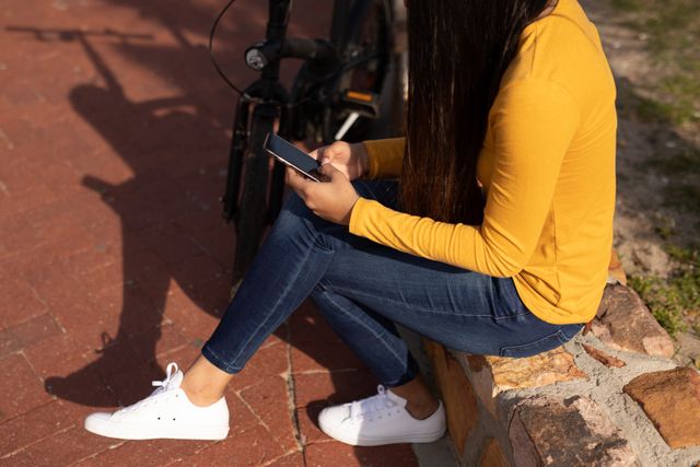 This image depicts a woman sitting outdoors on a sunny day, using her smartphone next to a bicycle. She is dressed casually in jeans and a yellow shirt, enjoying her free time in nature. This image can be used for promoting outdoor activities, leisure time, technology use, or lifestyle content.