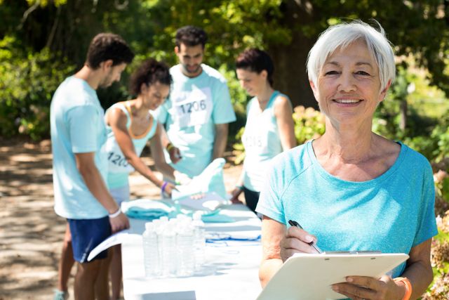 Senior woman coach smiling while athletes are registering for a marathon in a park. The coach holds a clipboard, and the athletes are gathered around a table with registration materials and water bottles. This image can be used for promoting community events, sports organizations, fitness programs, and volunteer activities.