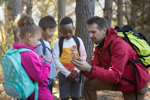 Teacher and children examining a plant in a jar during an outdoor educational activity. Ideal for use in educational materials, nature exploration themes, and promoting outdoor learning experiences.
