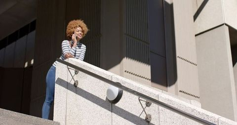 African American woman leaning on concrete railing talking on phone, brutalist backdrop