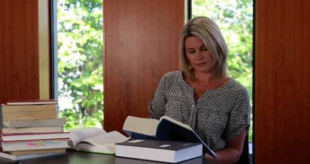 Blonde woman is reading book at table with a stack of books, embracing learning and education. She is seated in a room with large windows revealing a leafy outdoor view. Ideal for depicting education, academic research, studying, or literary pursuits. Could be used on websites, blogs, or posters about education, personal development, and libraries.