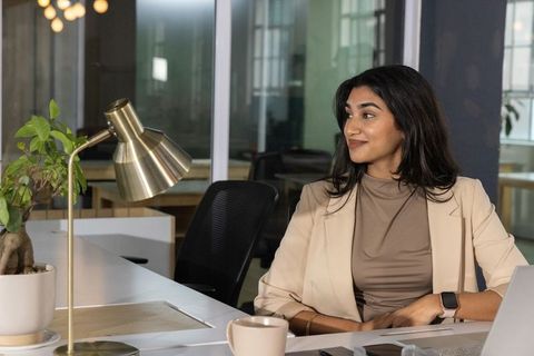 Confident indian businesswoman working at modern glass office desk with laptop and brass lamp