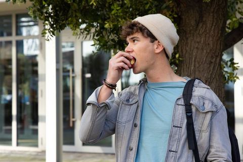 Teen biting red apple standing outside campus by tree wearing denim jacket and beanie