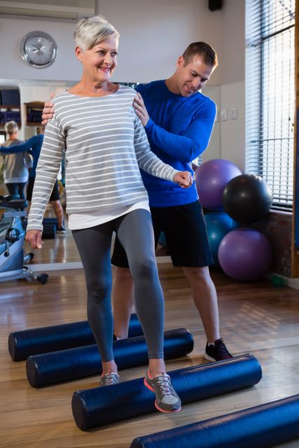Physiotherapist assisting senior woman in performing exercise on foam roll in clinic