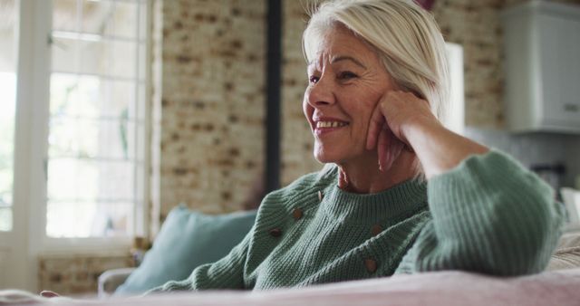 Elegant senior woman with gray hair relaxing on a couch in a living room, wearing a green sweater and smiling warmly. Bright, cozy living room in the background with natural light coming through the windows. Perfect for use in articles about aging, living healthy, comfortable home environments, and senior lifestyle.