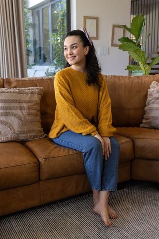 Smiling woman sitting on caramel leather sofa wearing mustard sweater in cozy living room