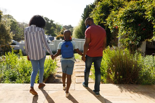 African-American parents walking with their son who is wearing a school backpack. This image can be used for themes related to family bonding, education, back to school, parenting, and outdoor activities. Ideal for advertisements, educational materials, and family-oriented content.