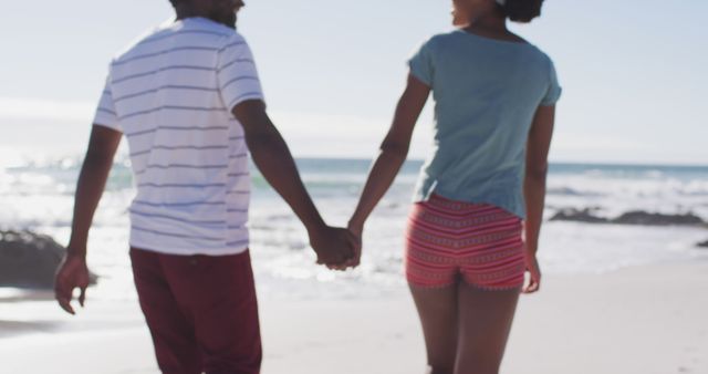 Romantic couple holding hands and walking along a sunny beach. This image can be used for travel advertisements, relationship blogs, summer vacation promotions, and social media posts emphasizing romance and tropical destinations.