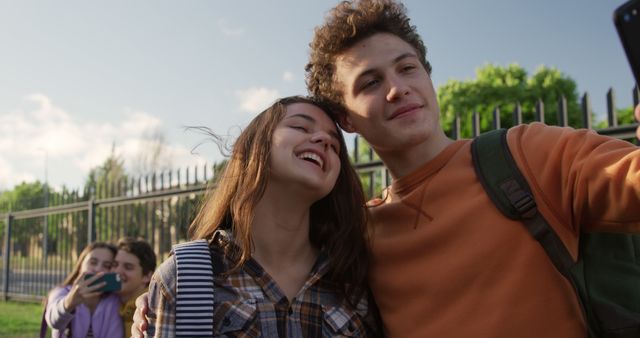 Happy caucasian teenage girl and boy taking selfie embracing outside school on sunny afternoon. Communication, social media, school, adolescence, romance, friendship, summer and education, unaltered.
