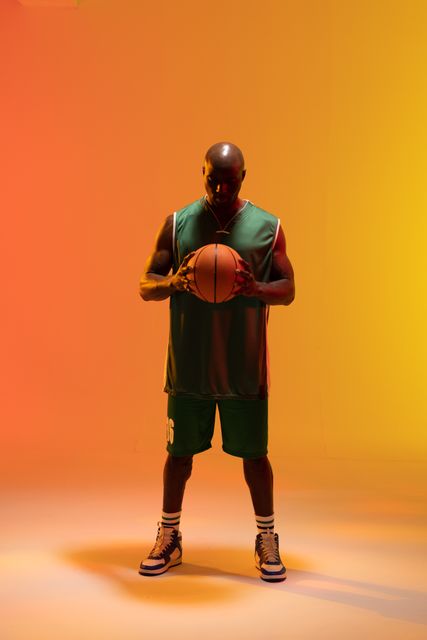 African American male basketball player standing confidently while holding a basketball in a studio with orange lighting. Ideal for use in sports promotions, fitness campaigns, advertisements for athletic wear, and motivational posters.